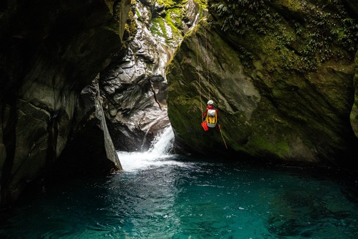 Person rappelling beside a waterfall into a rocky canyon with clear blue water.
