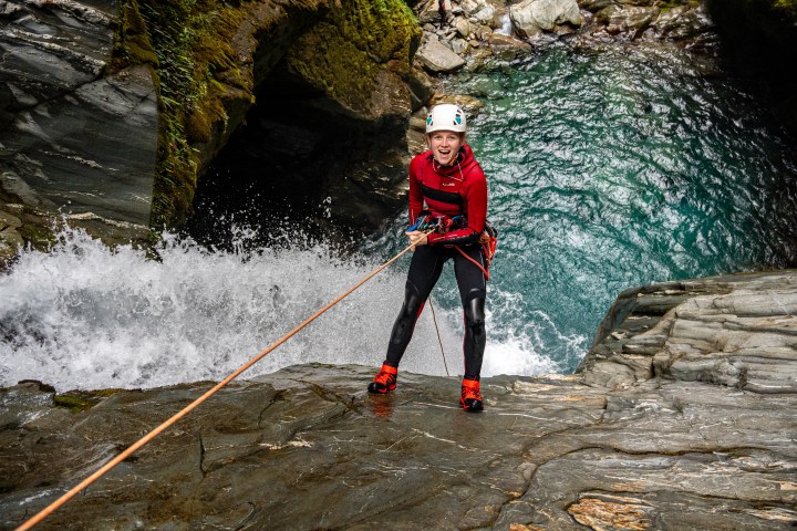 Person rappelling down rocky waterfall, wearing helmet and red jacket.
