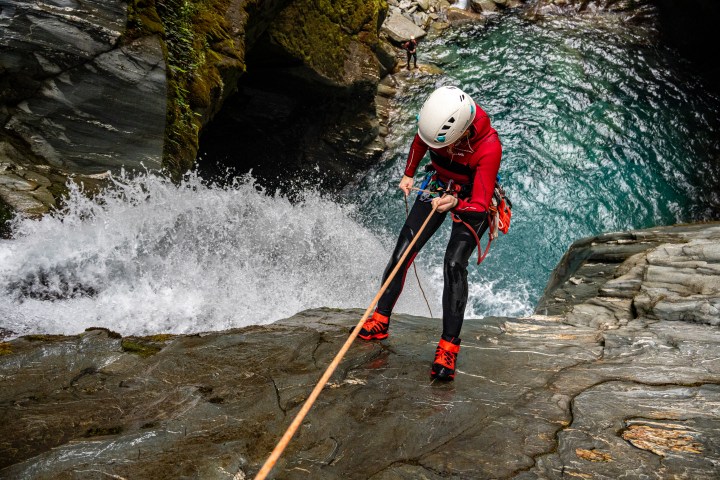Person canyoning down a waterfall with ropes and helmet beside a rocky pool.