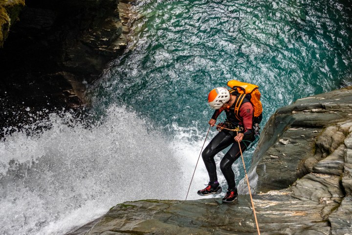 Person canyoneering down wet rocky cliff beside waterfall into a pool.