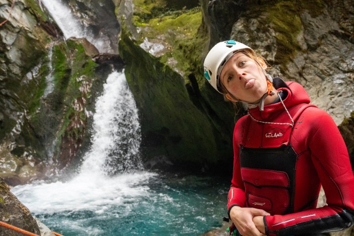 Person in red wetsuit and helmet poses playfully by waterfall.