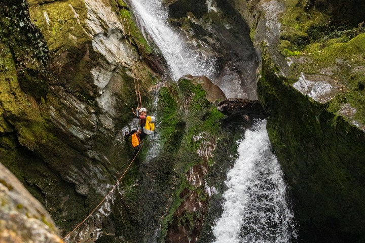 Person rappelling down a mossy cliff beside a waterfall in a lush canyon.