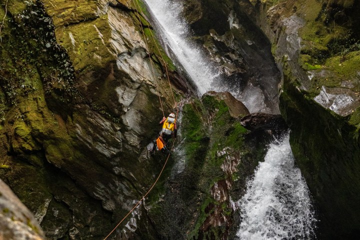 Person rappelling down a waterfall in a lush, rocky gorge.