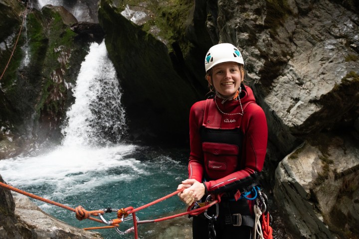 Person in red wetsuit and helmet smiling near waterfall with climbing gear.