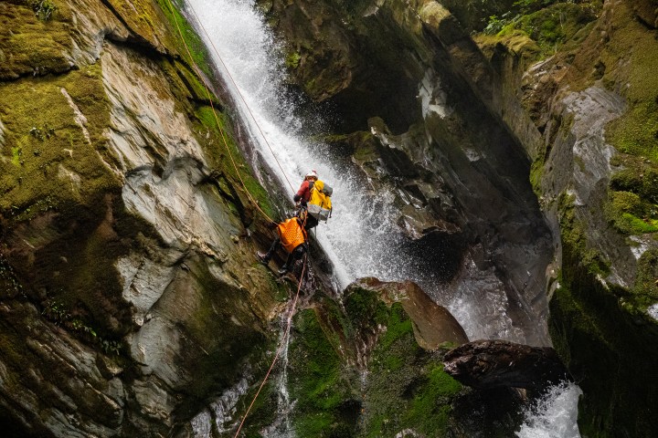Two people canyoning down a steep mossy rock face with rushing waterfall.