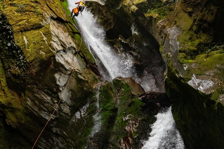 Person rappelling down a waterfall in a rocky canyon with lush greenery.