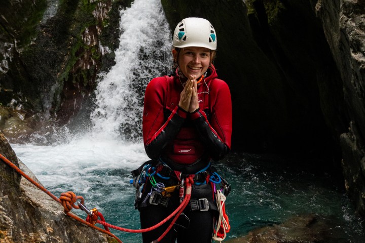 Person in red wetsuit and helmet stands by a waterfall in a rocky canyon.