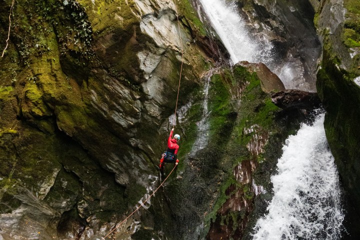 Person in red gear rappelling down a waterfall in a rocky gorge.