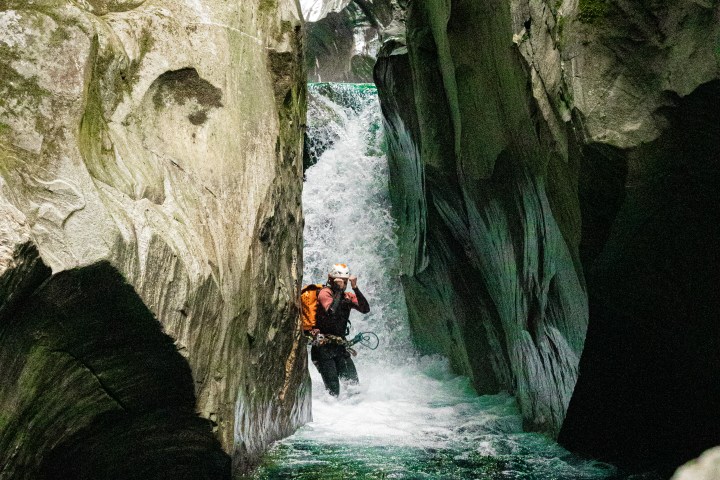 Person canyoning in a narrow, mossy gorge with waterfalls and blue water.
