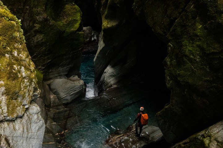 Person with orange backpack near flowing river in rocky gorge.