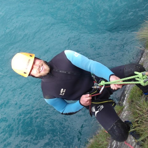 Person in wetsuit climbing cliff with ropes above blue water.