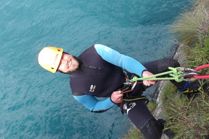 Person in wetsuit climbing cliff with ropes above blue water.
