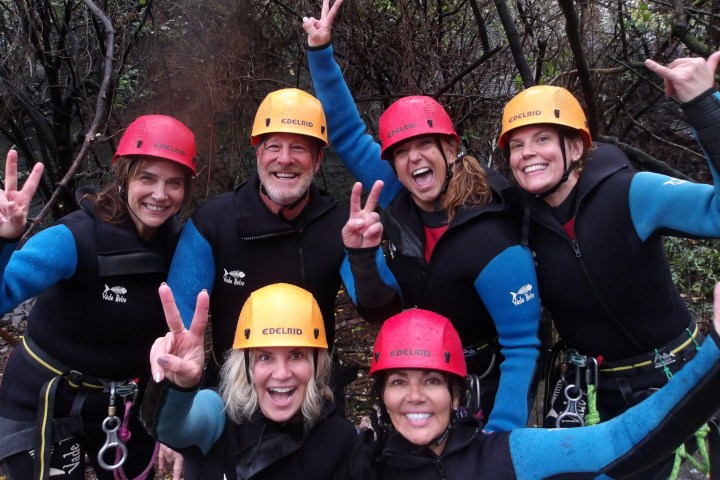 Six people in wet suits and helmets smiling and making peace signs in a forest setting.