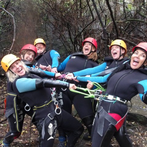 Group in wetsuits and helmets laughing, linked by ropes in a forest setting.