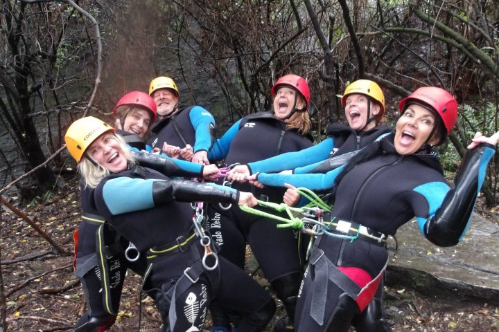 Group in wetsuits and helmets laughing, linked by ropes in a forest setting.