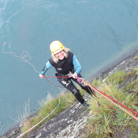 Person in a helmet and wetsuit climbing with ropes beside a body of water.