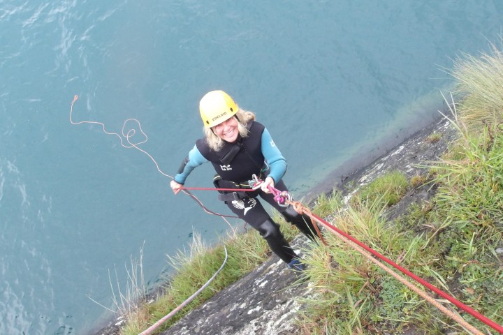 Person in a helmet and wetsuit climbing with ropes beside a body of water.