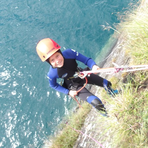 Person in helmet and wetsuit rappelling down a cliff with water below.