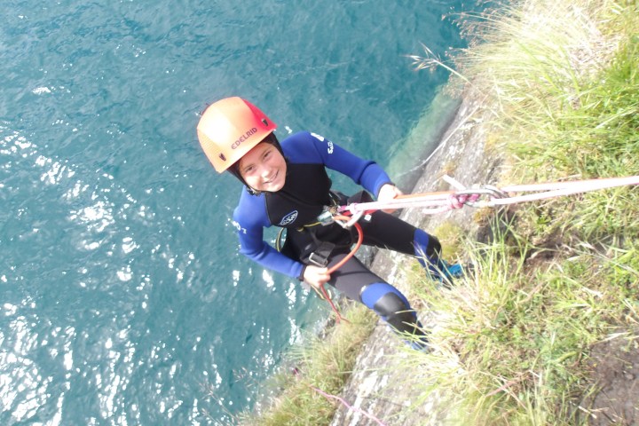 Person in helmet and wetsuit rappelling down a cliff with water below.