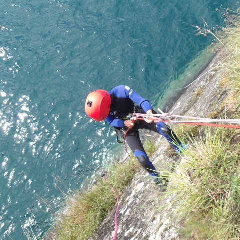 Person in wetsuit and helmet abseiling down a cliff towards a body of water.