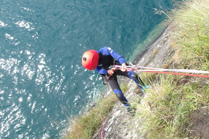 Person in wetsuit and helmet abseiling down a cliff towards a body of water.