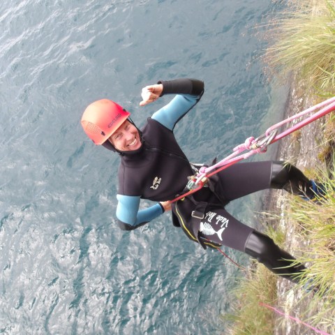 Person in wetsuit and helmet rappelling beside a body of water, smiling and gesturing with a thumb up.