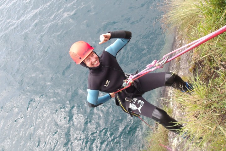 Person in wetsuit and helmet rappelling beside a body of water, smiling and gesturing with a thumb up.