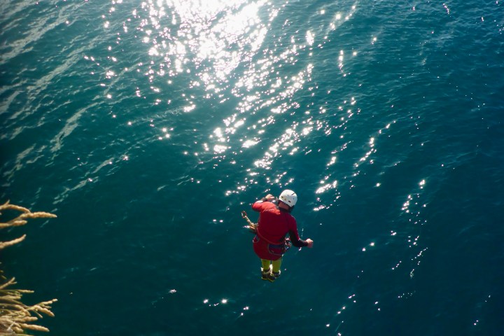 Person in red gear jumping into blue water from cliff, sunlight reflecting off surface.