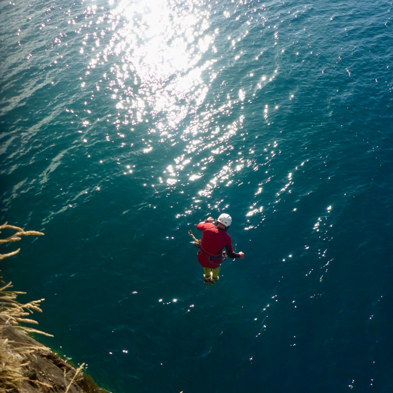 Person in red gear jumping into blue water from cliff, sunlight reflecting off surface.