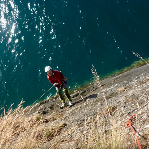 Person in climbing gear rappelling down a cliff with water below and grass on the cliff.