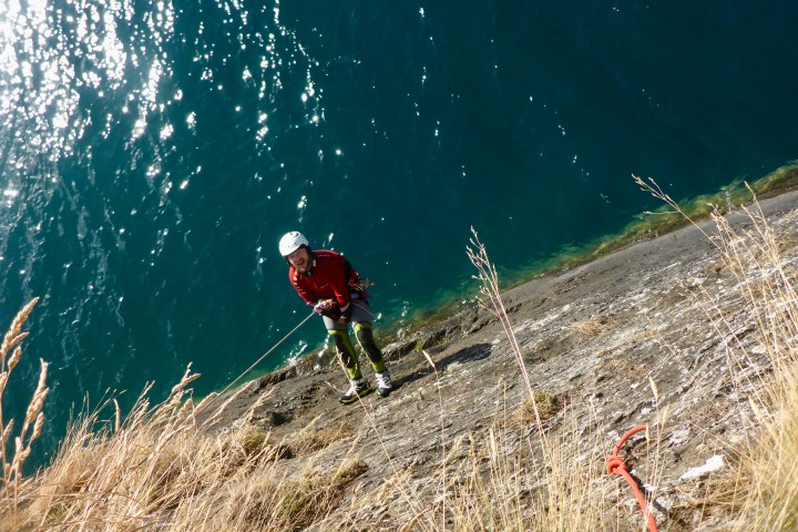 Person in climbing gear rappelling down a cliff with water below and grass on the cliff.