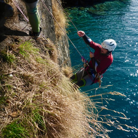 Person abseiling down a steep cliff face near a body of water, wearing a helmet and climbing gear.