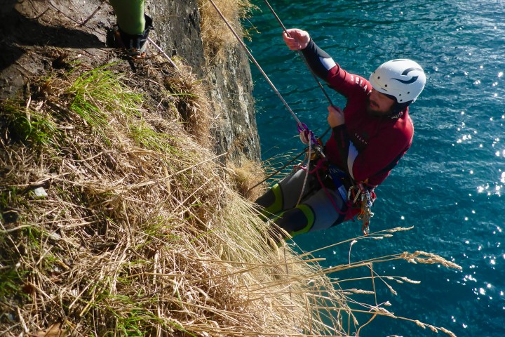 Person abseiling down a steep cliff face near a body of water, wearing a helmet and climbing gear.