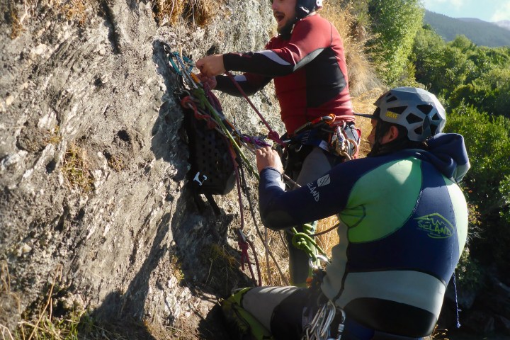 Two climbers in wetsuits and helmets securing ropes on a rocky cliffside with greenery around.