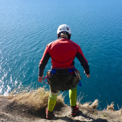 Person in wetsuit and helmet preparing to jump off cliff into water.