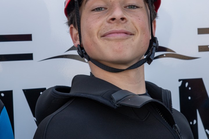 Person in a blue wetsuit and red helmet smiling in front of a sign.