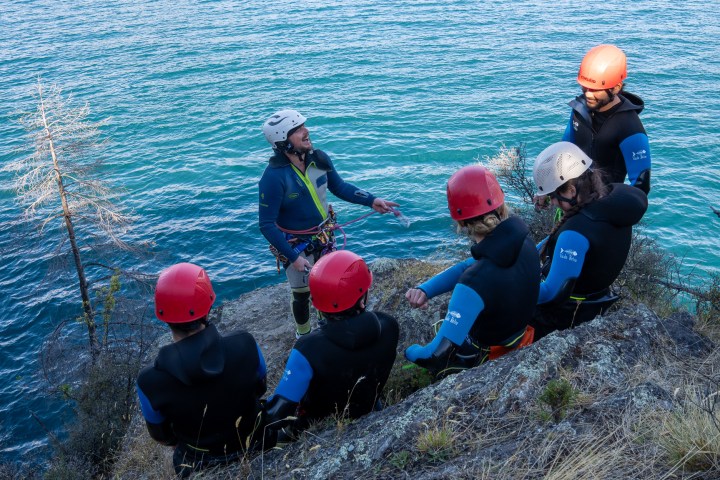 Group of people in helmets and wetsuits listening to an instructor by a body of water.