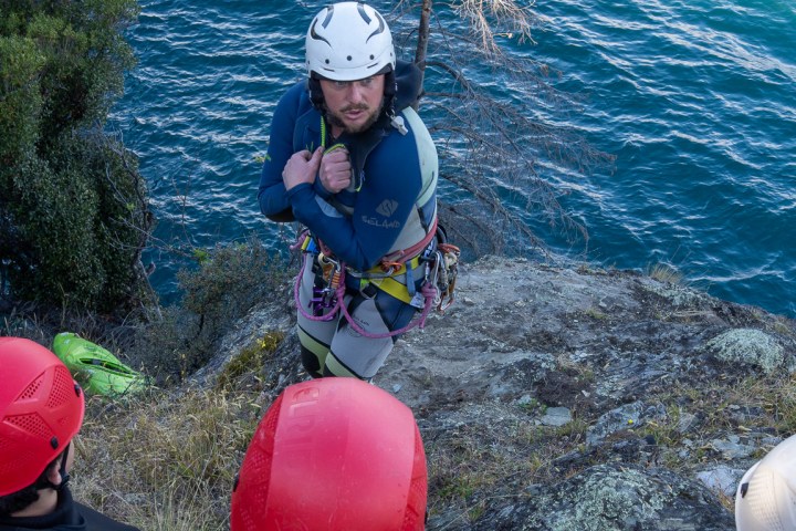 Group with helmets watching instructor near cliff by water.