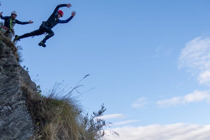 Person in mid-air jumping from cliff over water with hills in the background.