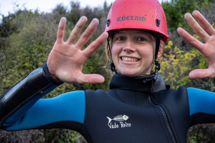 Person in wetsuit and helmet smiling with raised hands outdoors.