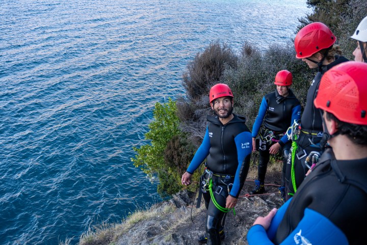 Group of people in helmets and wetsuits on a cliff by the water.
