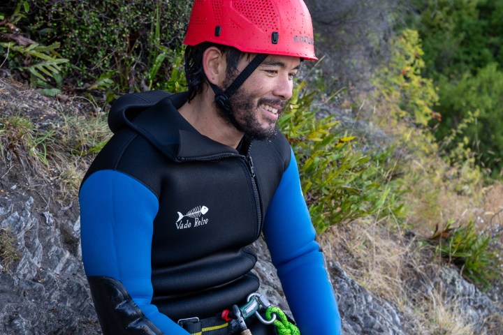 Person wearing a red helmet and wetsuit sitting on a rocky surface.