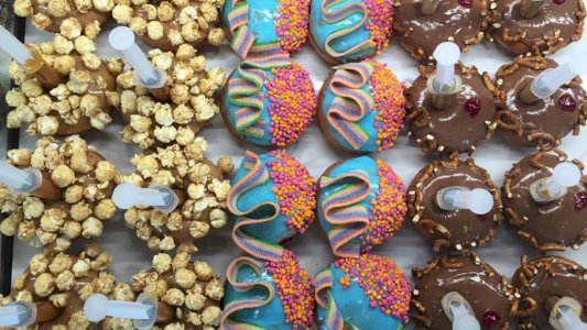 Three rows of colorful decorated donuts with syringes, topped with popcorn, sprinkles, and pretzels.
