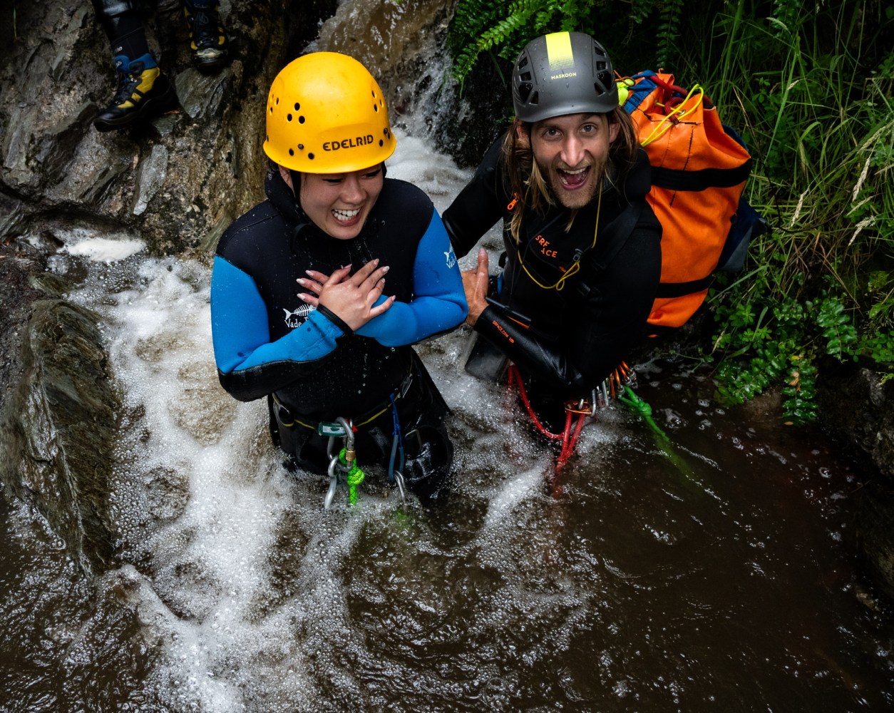 Two people in wetsuits and helmets smiling while standing in a rocky stream.