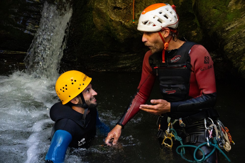 Two people with helmets and wetsuits standing in water near a waterfall, one talking to the other.