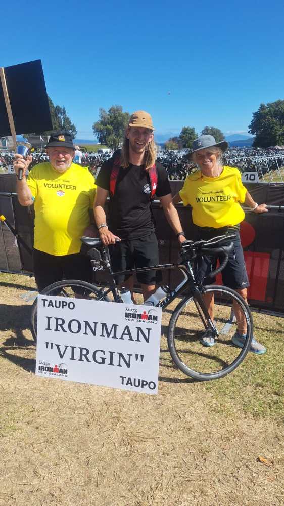 Three people with a bicycle and Ironman sign outdoors, two wear yellow volunteer shirts.
