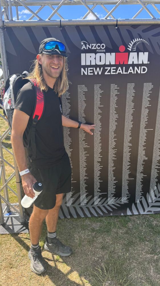Person pointing to names on Ironman New Zealand board outdoors, wearing black outfit and cap with sunglasses.