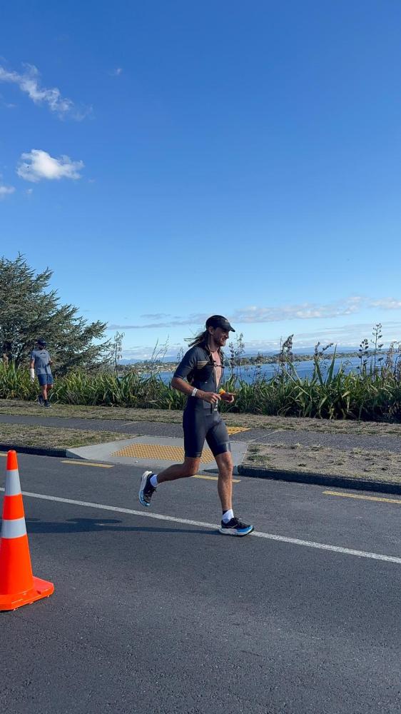 Runner in a triathlon suit on a paved road by the seashore under clear blue sky.