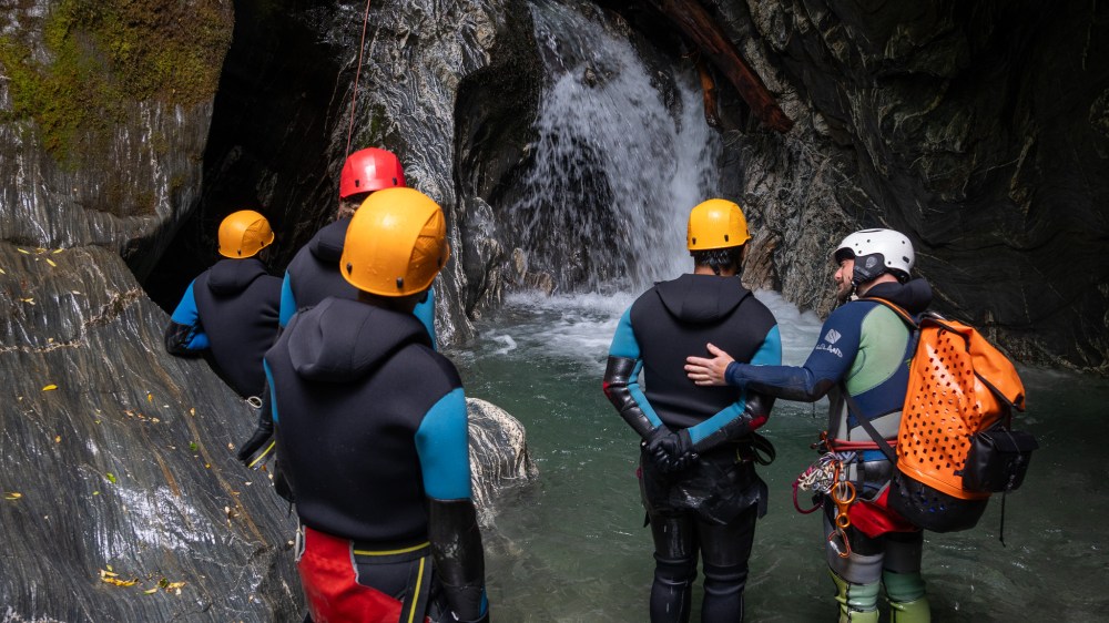 Group of people in wetsuits and helmets canyoning near a waterfall.