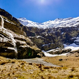 Mountain landscape with snow, waterfall, and rocky terrain under a clear blue sky.